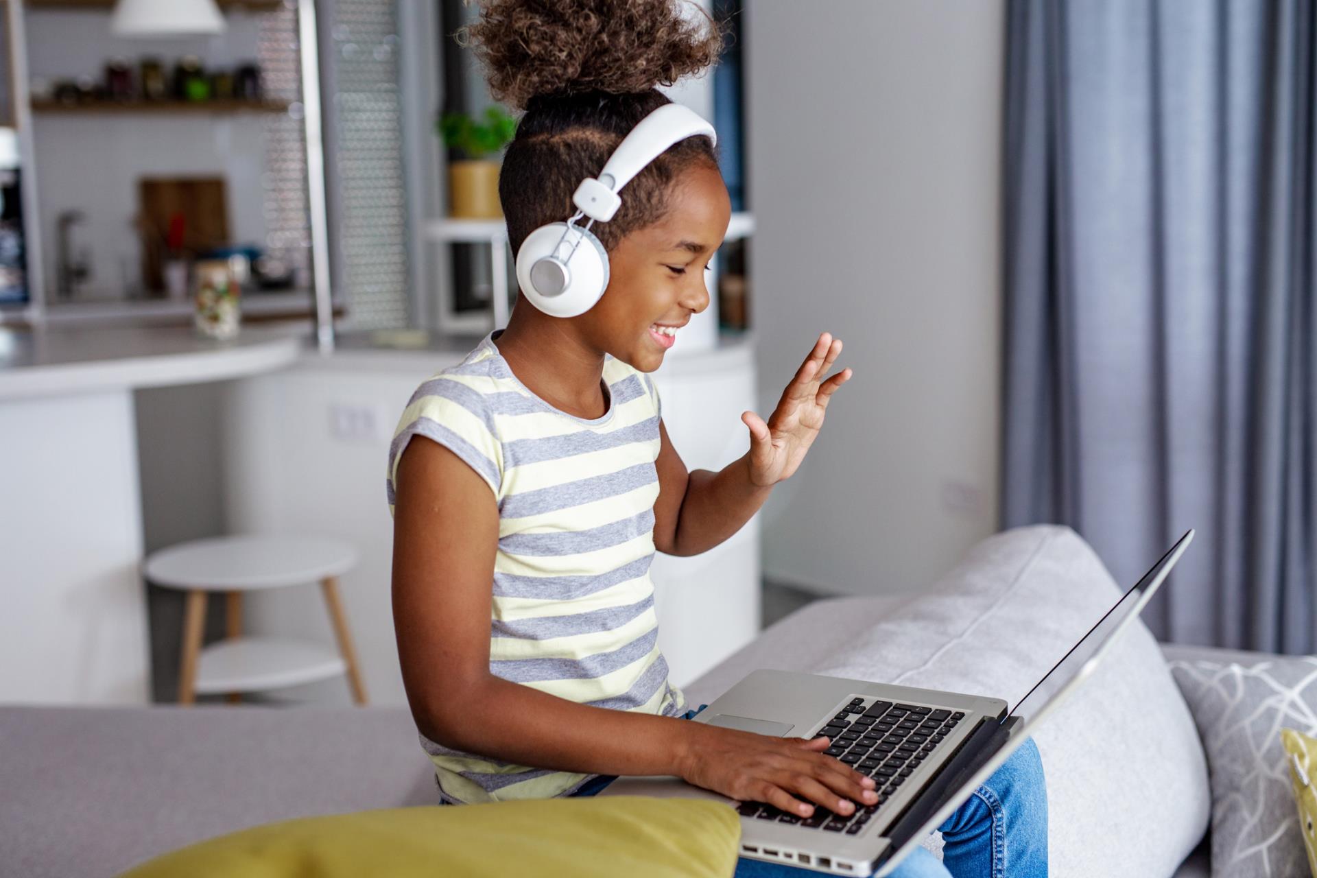 Student at home wearing headphones and waving at laptop in her lap